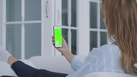 Woman-using-mobile-phone-vertical-green-screen.-Female-holding-in-hand-portable-gadget-close-up-indoors-home-living-room.-Mock-up-for-tracking-or-watching-content.-Blurred-room-on-the-background
